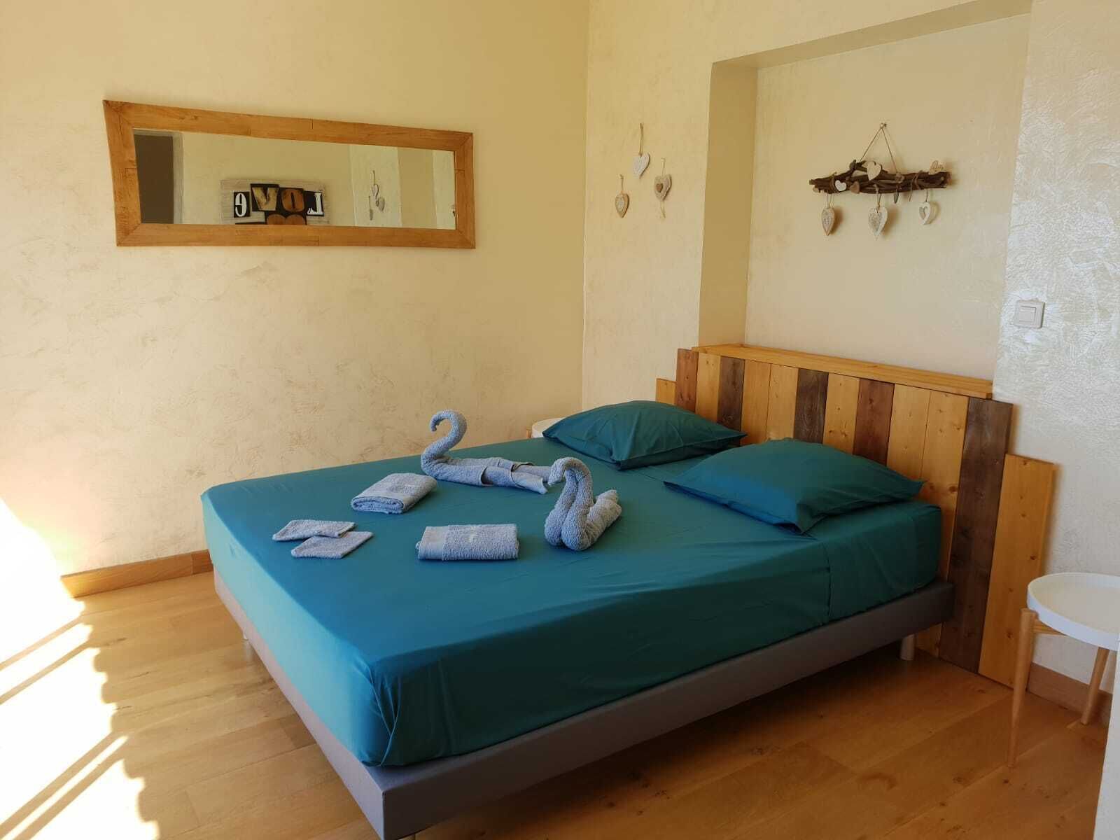 Una cama individual con cabecera de madera, ropa de cama color turquesa y dos almohadas. Un espejo cuelga sobre la cama y se ve una decoración de corazones en la pared.