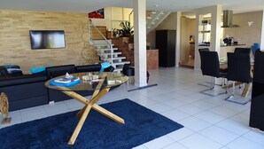Ein modernes Wohnzimmer mit einem schwarzen Sofa, einem Glastisch und einem blauen Teppich. Eine Treppe führt zu einer Küchenzeile mit Esstisch und Stühlen.