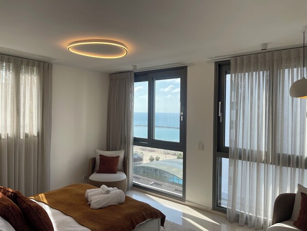 A modern hotel room with a large bed, a seating area with a chair and a small table, a balcony with a view of the sea, and a ceiling light fixture. - Stunning beachfront duplex penthouse with infinity rooftop pool & parking (Tel Aviv-Yafo)