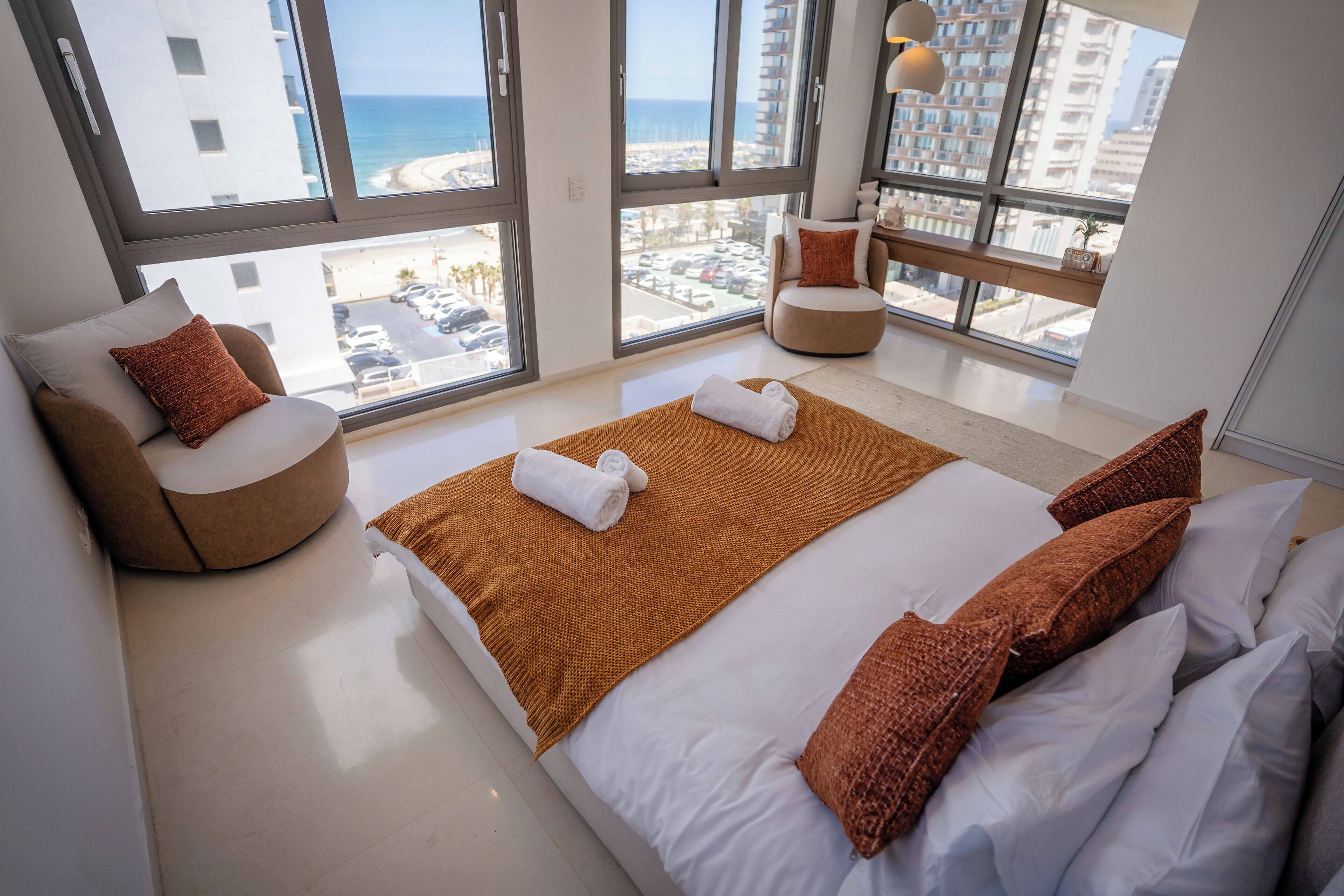 A modern hotel room with a large bed, a round armchair, and a view of the beach and cityscape through large windows.