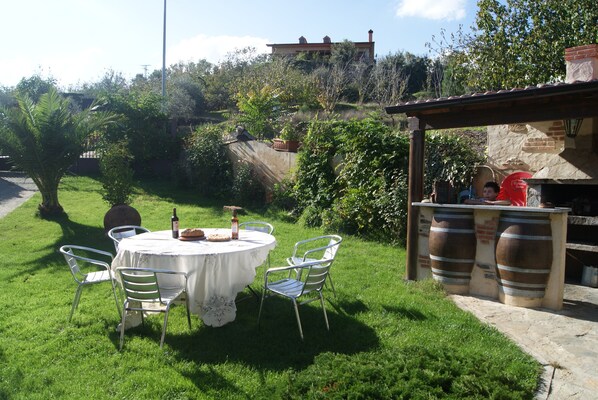 A round table with chairs and a wine bottle, set on a grassy lawn with a stone wall and a brick oven in the background. - Tranquility, friendship and good times. (Sotoserrano)