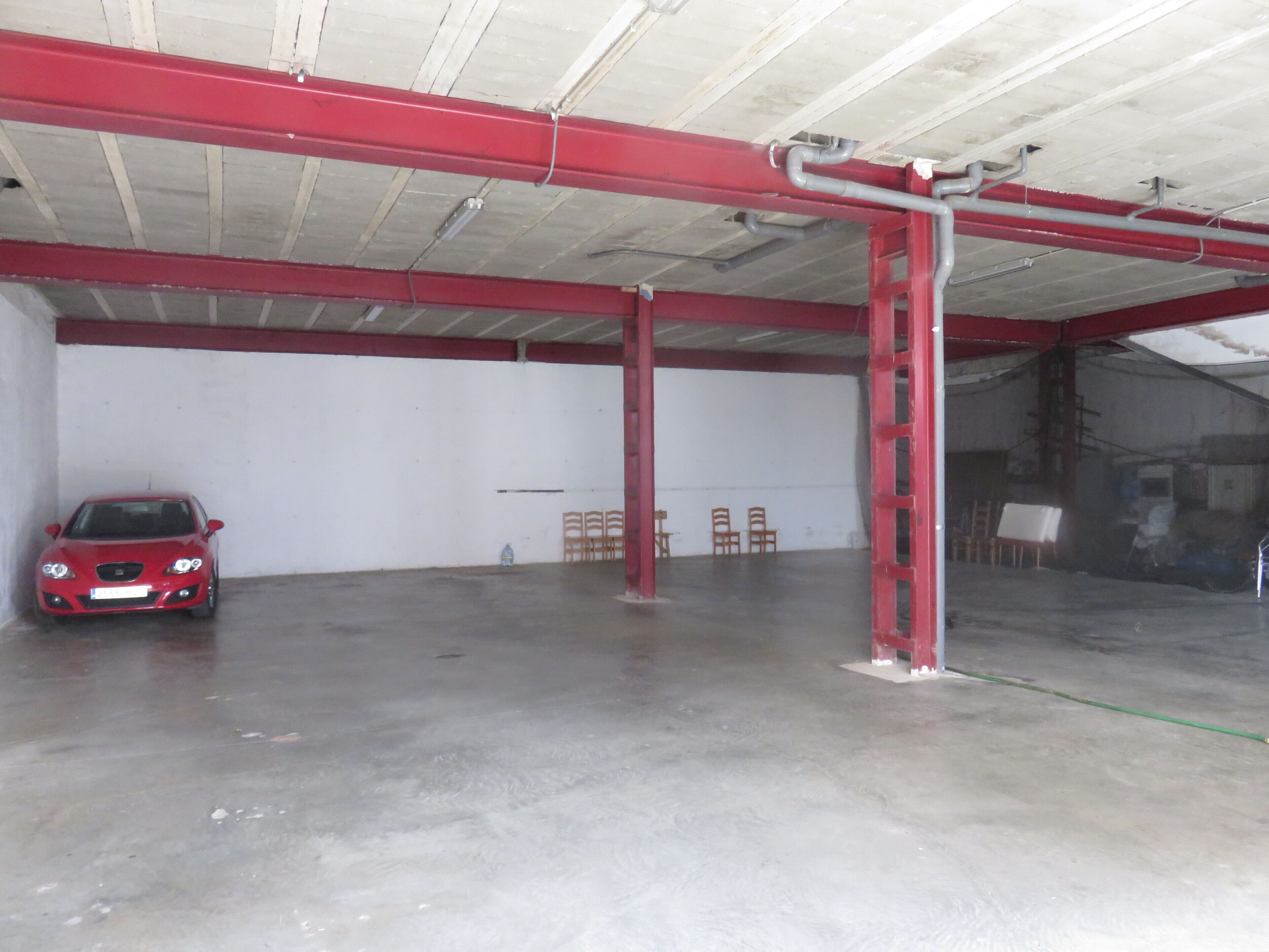 A spacious indoor area with a red car parked in the foreground, a white wall, and a concrete floor.