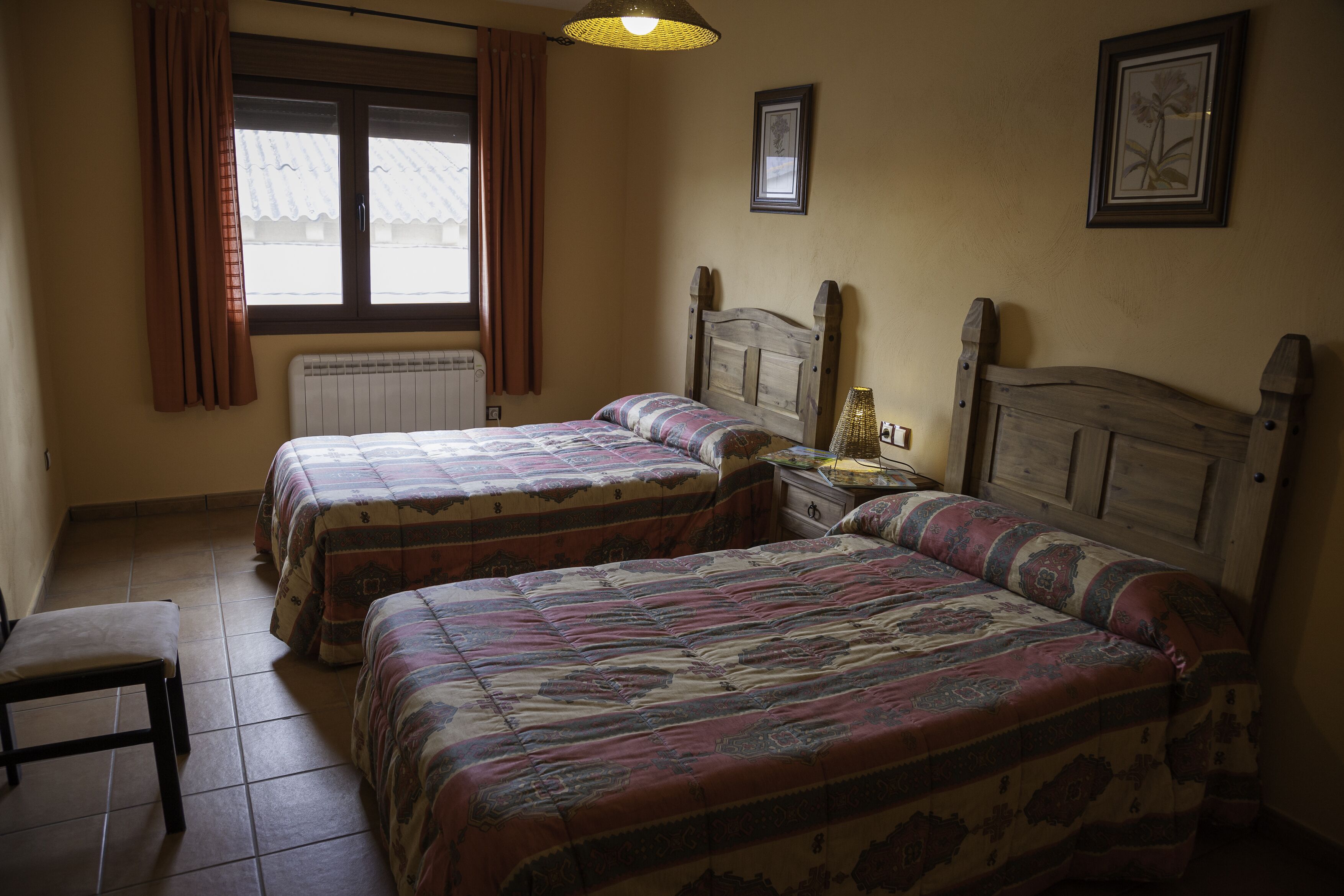 A bedroom with two beds, a window with curtains, a radiator, and a small table with a lamp.