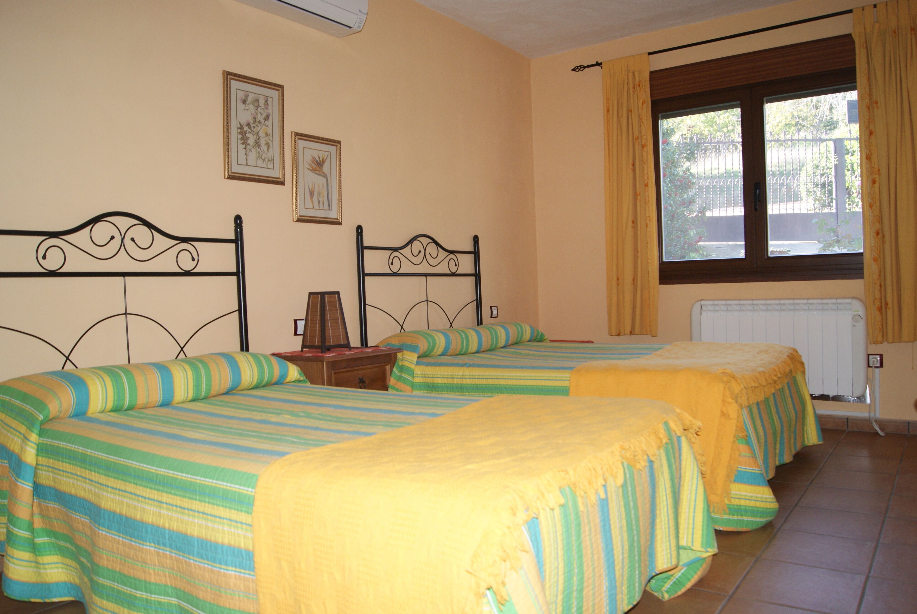 Una habitación con dos camas, cada una con ropa de cama a rayas y una colcha amarilla. Hay cuadros enmarcados en la pared, una ventana con cortinas y un radiador.