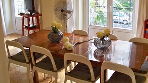 Un área de comedor con una mesa de madera, sillas blancas, un televisor sobre un mueble, un ventilador y vistas al exterior.