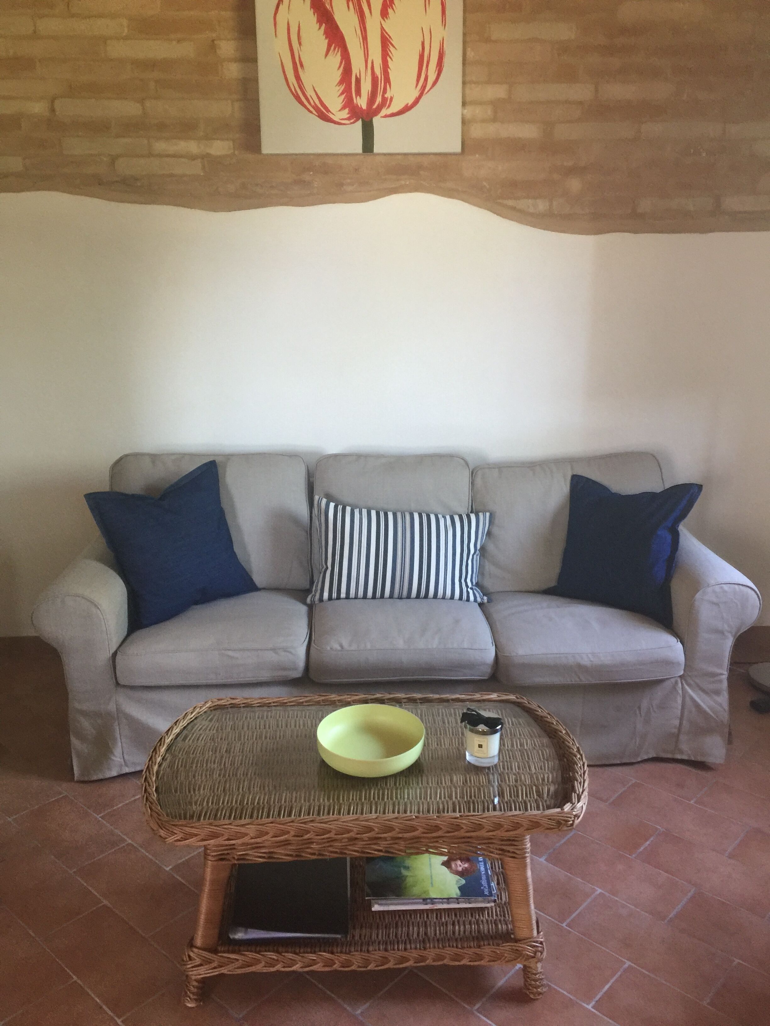Un salon avec un canapé gris, deux coussins bleus, un coussin à rayures, un bol jaune, une petite bouteille et un livre posés sur une table en osier.