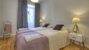 A double room with two beds, a window with curtains, bedside lamps, and a small bedside table with a lamp.