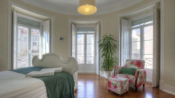 Un dormitorio con una cama, una silla, una planta en maceta y ventanas amplias.