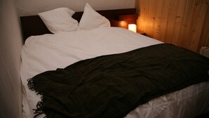 Un lit avec du linge de lit blanc et une couverture foncée, une table de chevet avec une bougie, et un lambris en bois sur le mur.