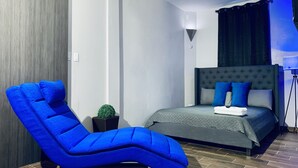 A modern bedroom with a tufted headboard bed, a blue cushioned chair, and a small potted plant. - Modern Suite - Pasavento Building (Aguadilla)