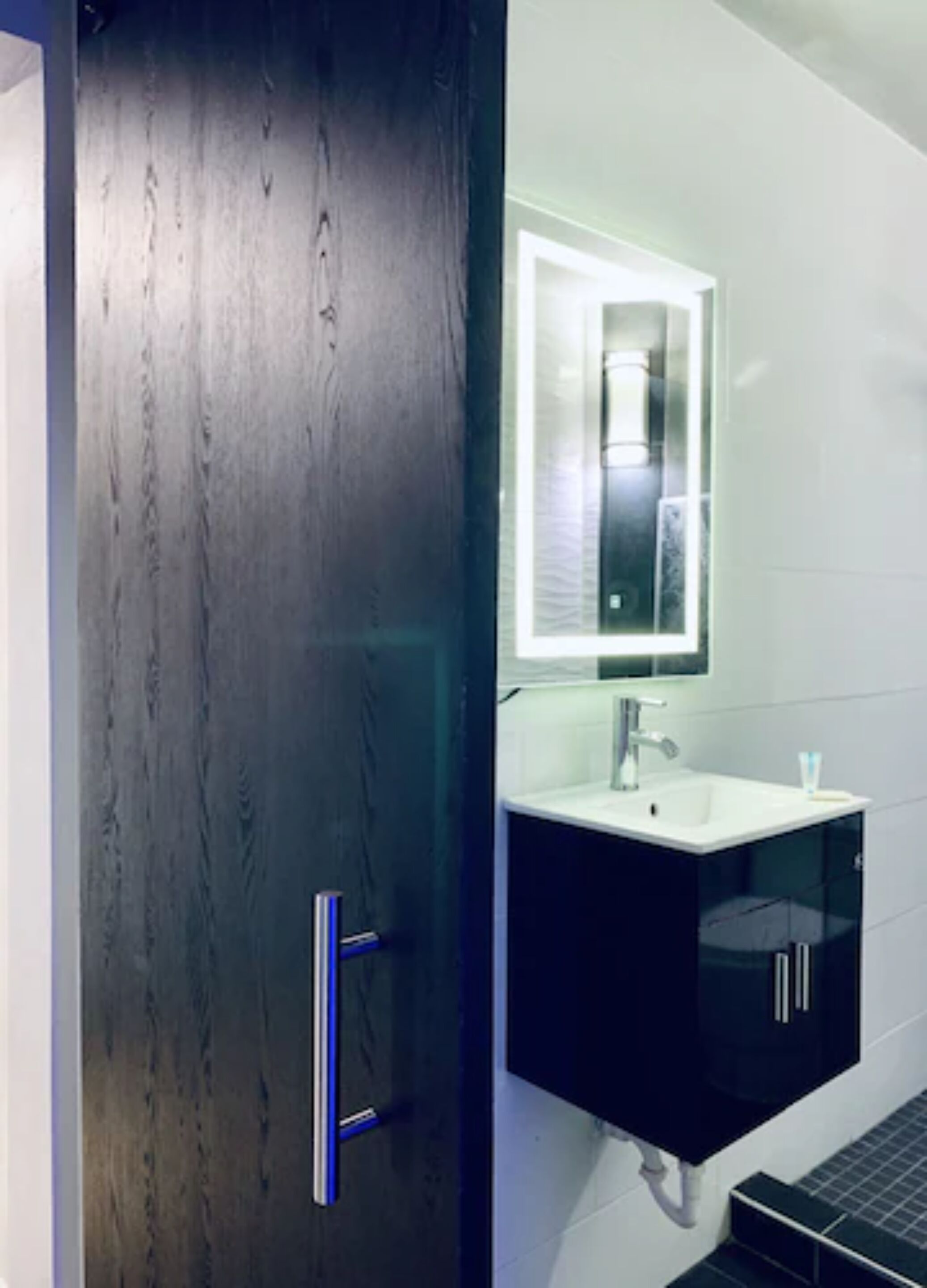 Une salle de bain moderne avec une porte en bois foncé, un lavabo blanc avec un miroir et des meubles noirs.