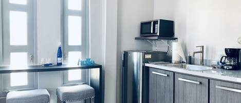 Une cuisine moderne équipée d’un réfrigérateur en acier inoxydable, d’un four à micro-ondes et d’une machine à café. On y trouve deux tabourets de bar et une petite table avec une bouteille et des verres.