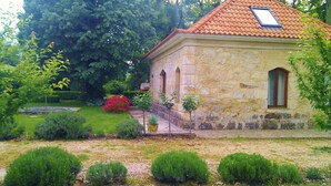 A stone building with a tiled roof, a small garden with shrubs and a flower bed, and a paved pathway leading to the entrance.