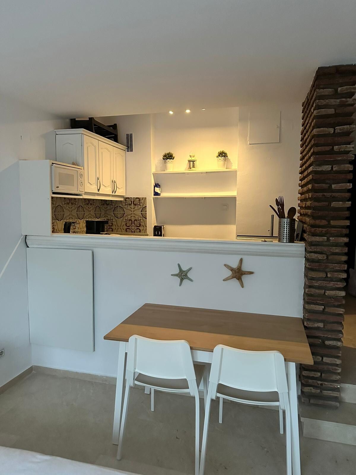 A compact kitchen with white cabinets, a microwave, a small island with potted plants, and a dining area with two white chairs.