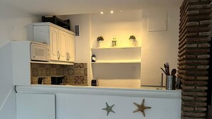 Une cuisine compacte avec des meubles blancs, une table à manger en bois et deux chaises blanches. On peut y admirer des étoiles de mer décoratives accrochées au mur, ainsi qu’un mur d’accent en briques.