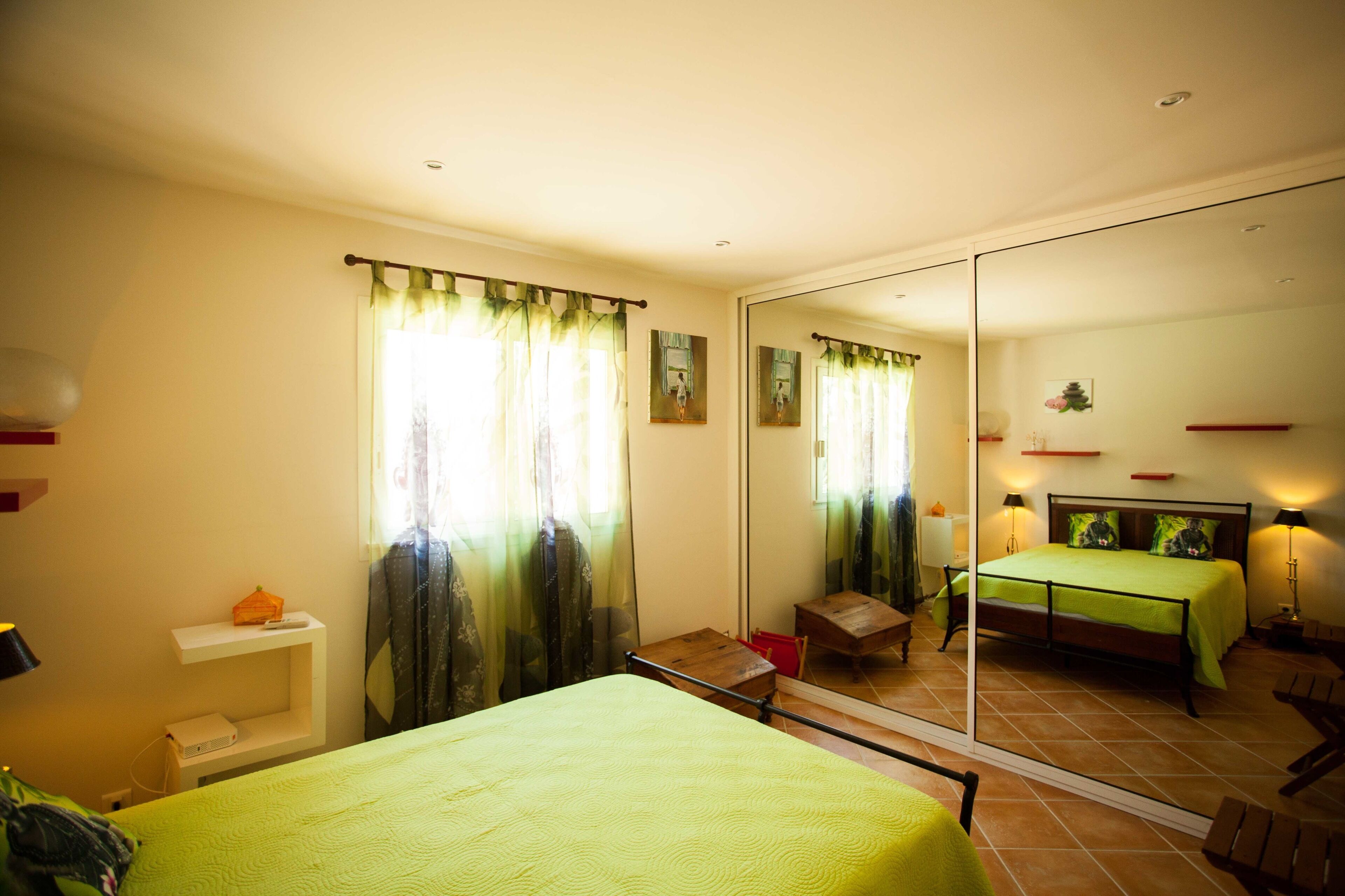 A bedroom with a bed, a chair, a small table, a mirror, and a window with curtains.