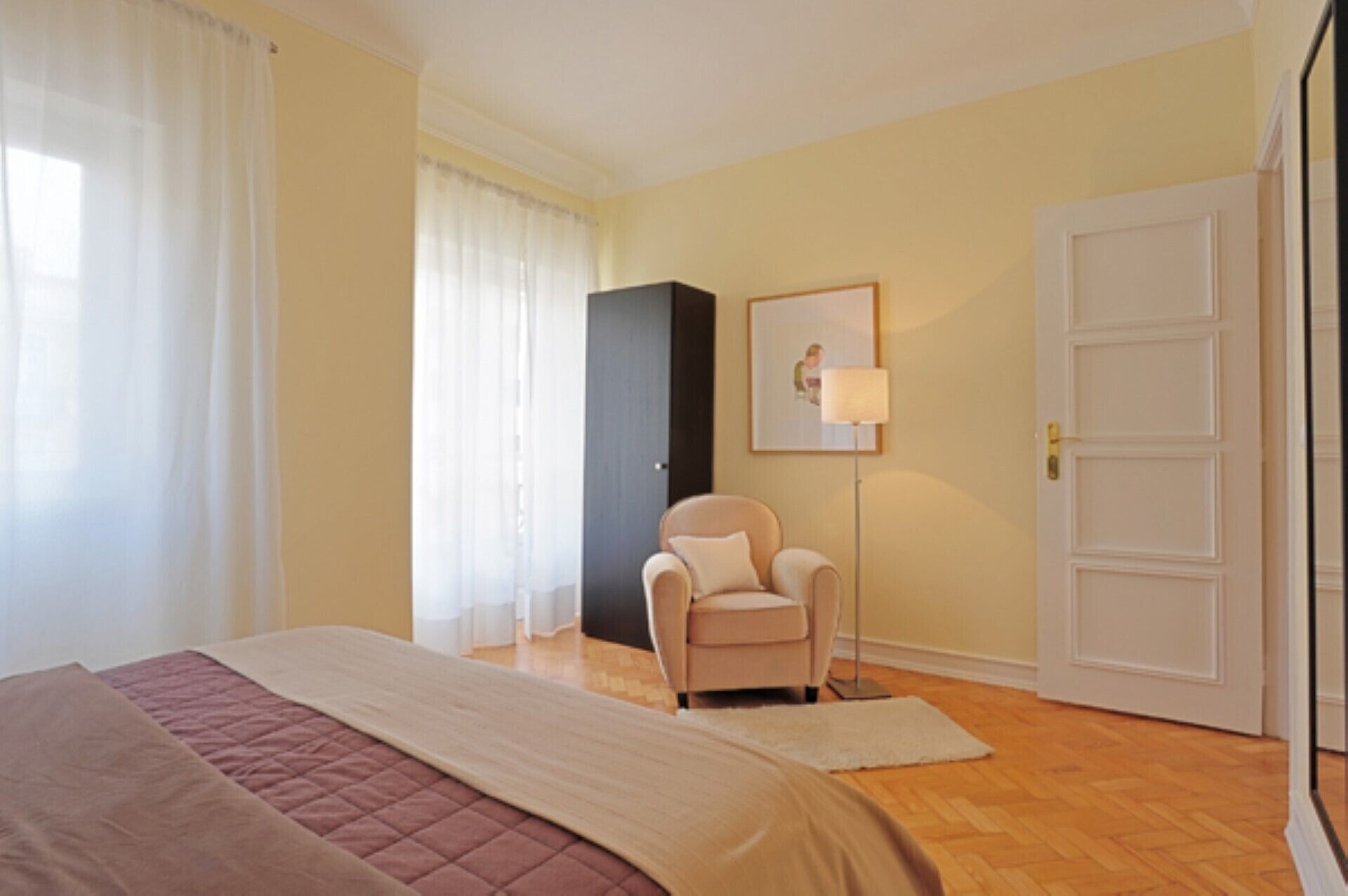 A hotel room with a bed, a chair, a desk, a lamp, and a door.