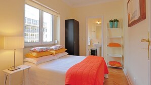 Une chambre à coucher comprenant un lit, une table de chevet, une fenêtre, une armoire, un miroir, une étagère avec des plantes et une porte.