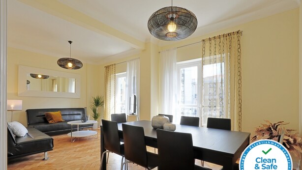 Un salon moderne avec un coin repas, comprenant un grand miroir, des canapés noirs, une table ronde et un luminaire suspendu.