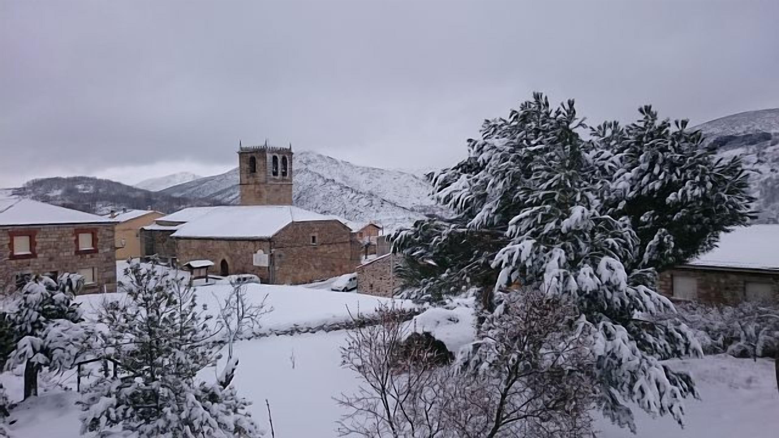 Uma aldeia nevada com edifícios de pedra, incluindo uma igreja com um campanário, rodeada por árvores e montanhas cobertas de neve.