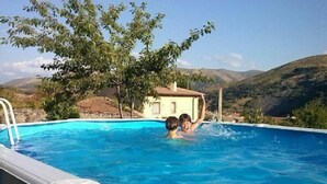 A swimming pool with two people in it, surrounded by a house and hills.