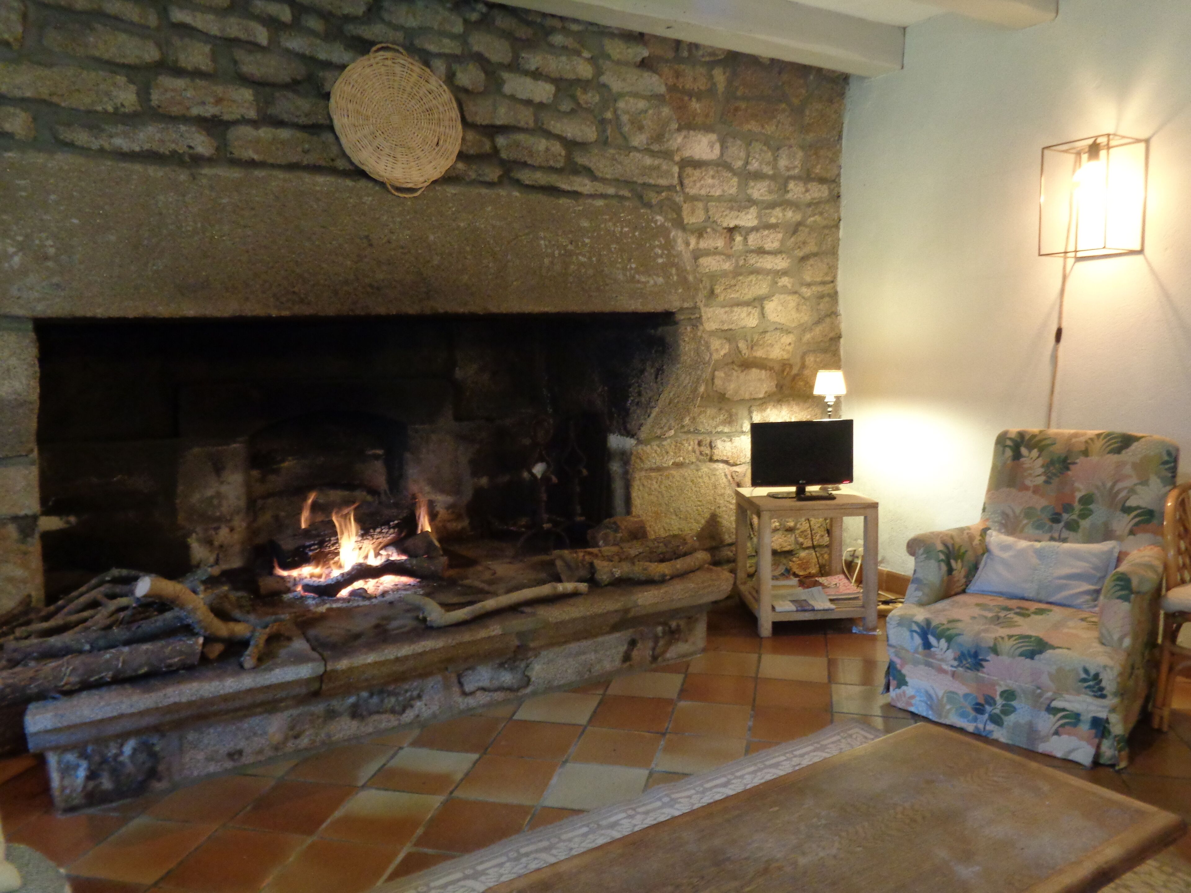 A stone fireplace with logs, a television on a wooden stand, a floral armchair, and a patterned rug on a tiled floor.