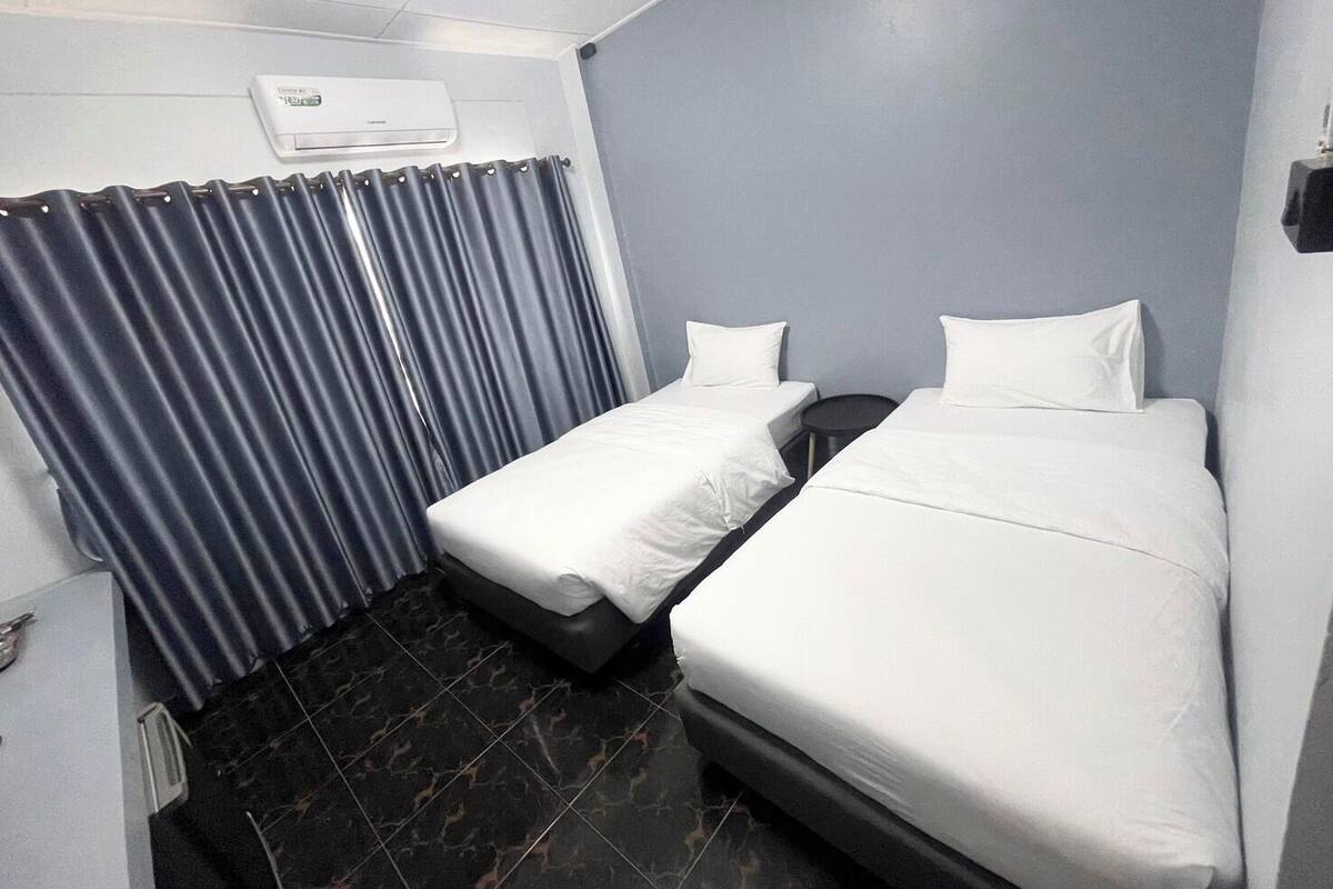 A hotel room with two single beds, a small table, a chair, a wardrobe, and an air conditioning unit.