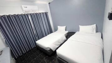 A hotel room with two single beds, a small table, a chair, a wardrobe, and an air conditioning unit.
