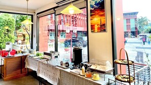 A breakfast buffet setup with a variety of food items and a coffee station, located in a room with large windows offering a view of the street outside. - Chainatgrand Hotel (Chai Nat)