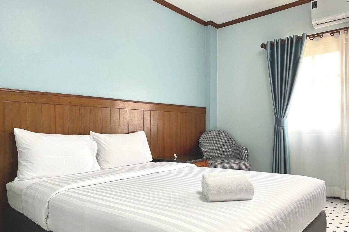 A hotel room with a large bed, a wooden headboard, a checkered chair, and a window with curtains.
