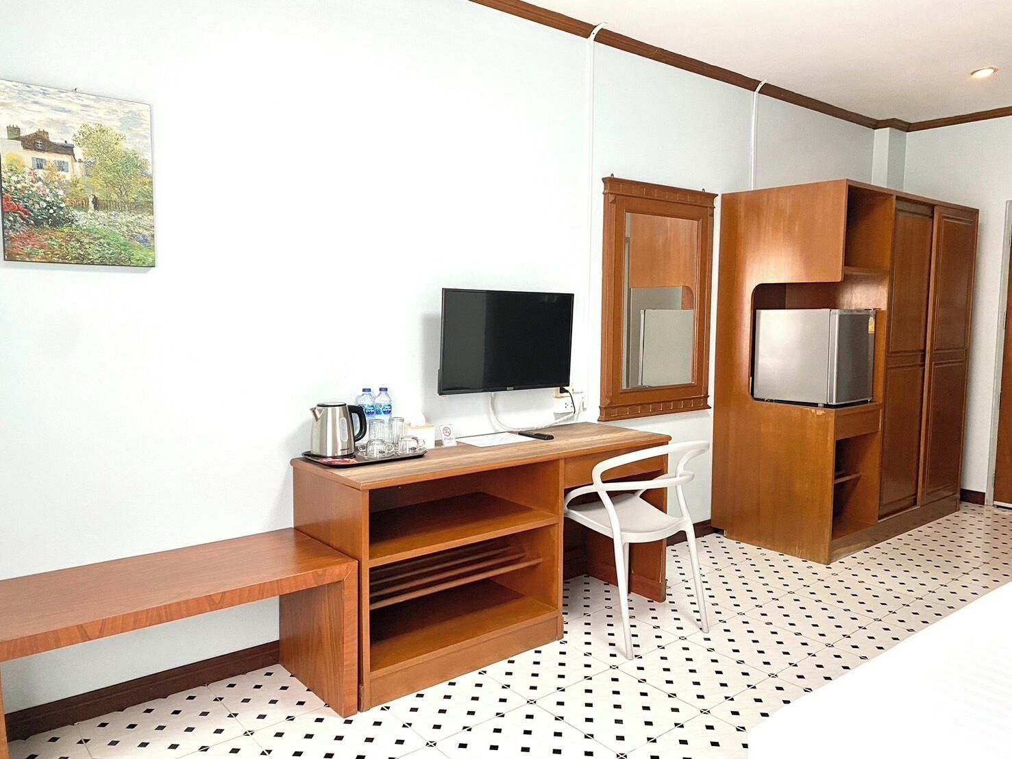 A compact hotel room with a wooden desk, a television, a refrigerator, and a white chair.