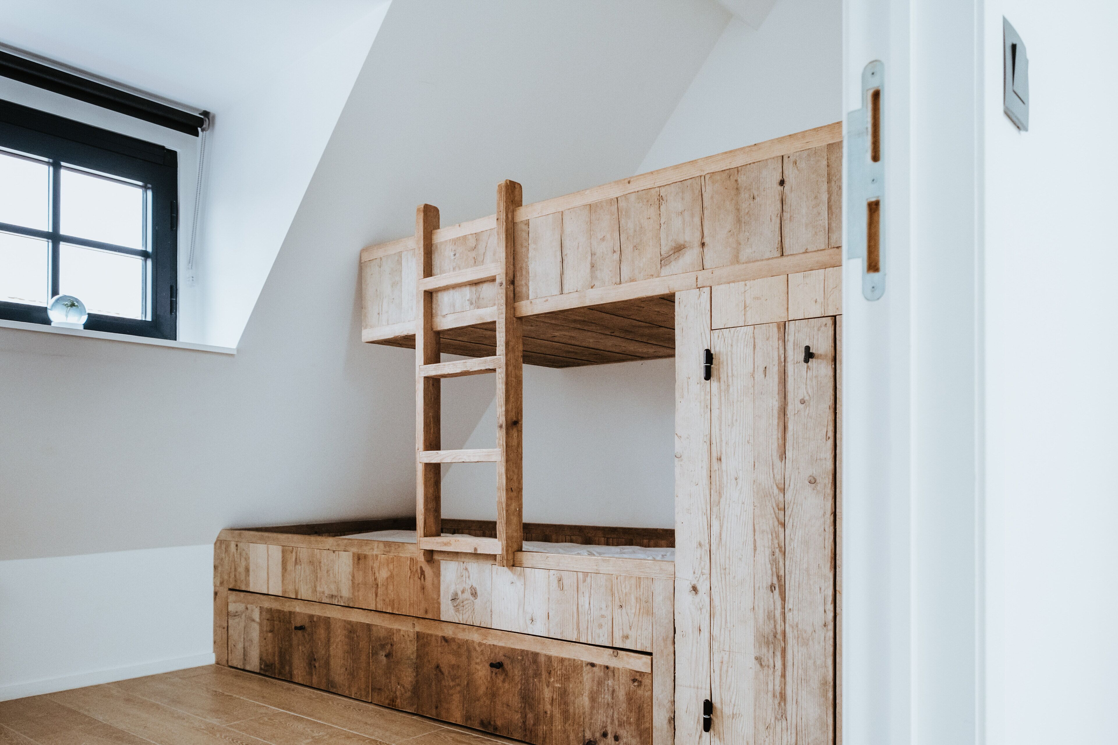 Una cama litera con escalera de madera, cajones integrados y una ventana con persianas.