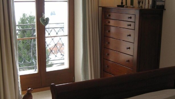 A bedroom with a bed, a dresser, a window with curtains, and a view of a balcony. - Luxury alpine apartment with warm indoor pool, sauna & fitness room (Gstaad)