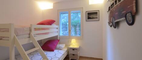 A child's bunk bed with a ladder, a bedside table with a lamp, a window with a view of greenery, and a wall-mounted decoration.