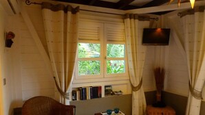 A cozy room with a wicker chair, a small table with glasses, a bookshelf, and a window with curtains. - Creole pavilion in the garden. (le francois)