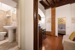Een badkamer met een wastafel, toilet en douche. Een houten deur leidt naar een woonruimte met een bank en een bed.