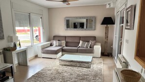 A modern living room with a sectional sofa, a coffee table, a rug, a ceiling fan, a large mirror, and a window with blinds. - Villa on Mar Menor Golf Resort, Murcia. High quality 3-bed villa with pool. (Torre-Pacheco)