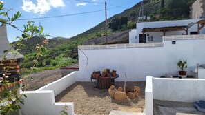 A patio area with a round table surrounded by potted plants, a white wall, and a view of a hillside. - Sandra's house  (Arona)
