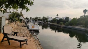 Eine Holzstele mit einer Liege und einem kleinen Tisch, ein Boot an der Anlegestelle, und ein ruhiges Gewässer mit Gebäuden im Hintergrund.