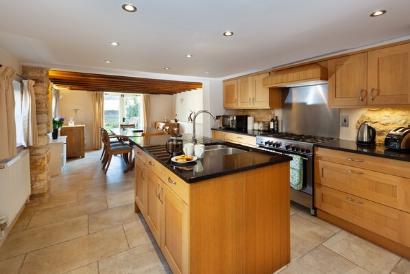 A modern kitchen with wooden cabinets, a central island, stainless steel appliances, and a dining area with natural light. - Camside, Chipping Campden - Taswell Retreats (Chipping Campden)