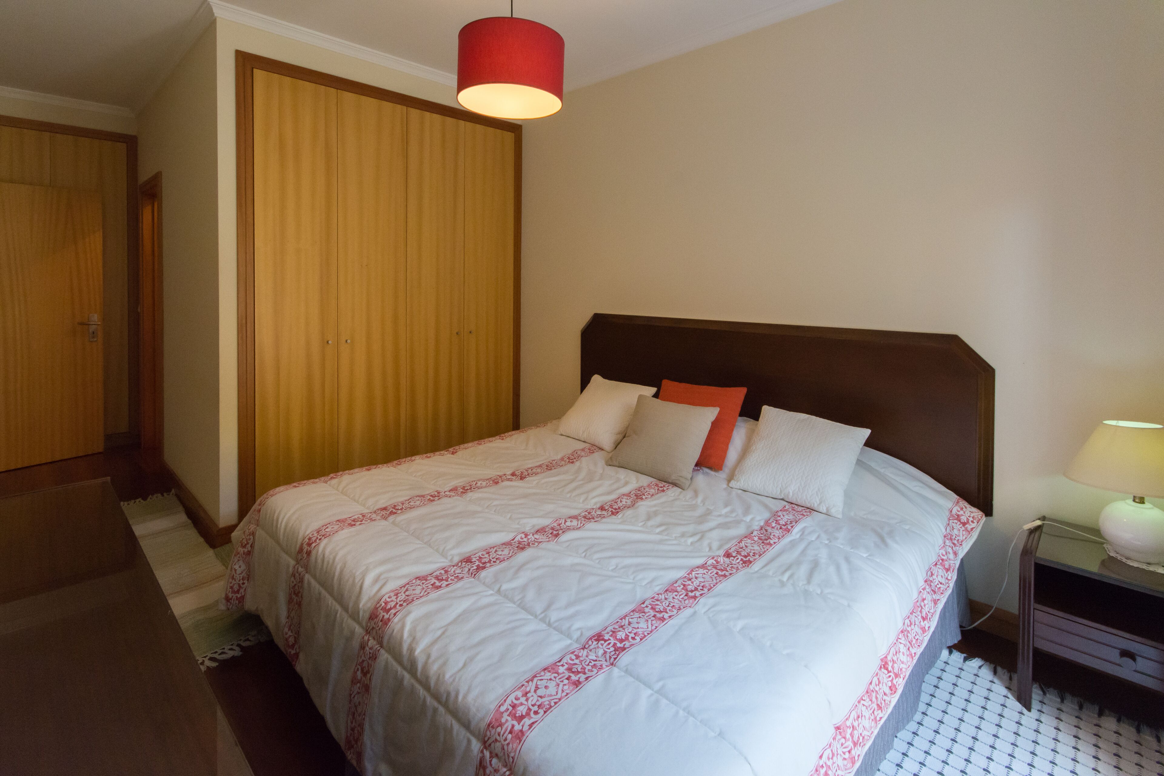 A neatly made bed with a white and red bedspread, a wooden wardrobe, and a nightstand with a lamp.