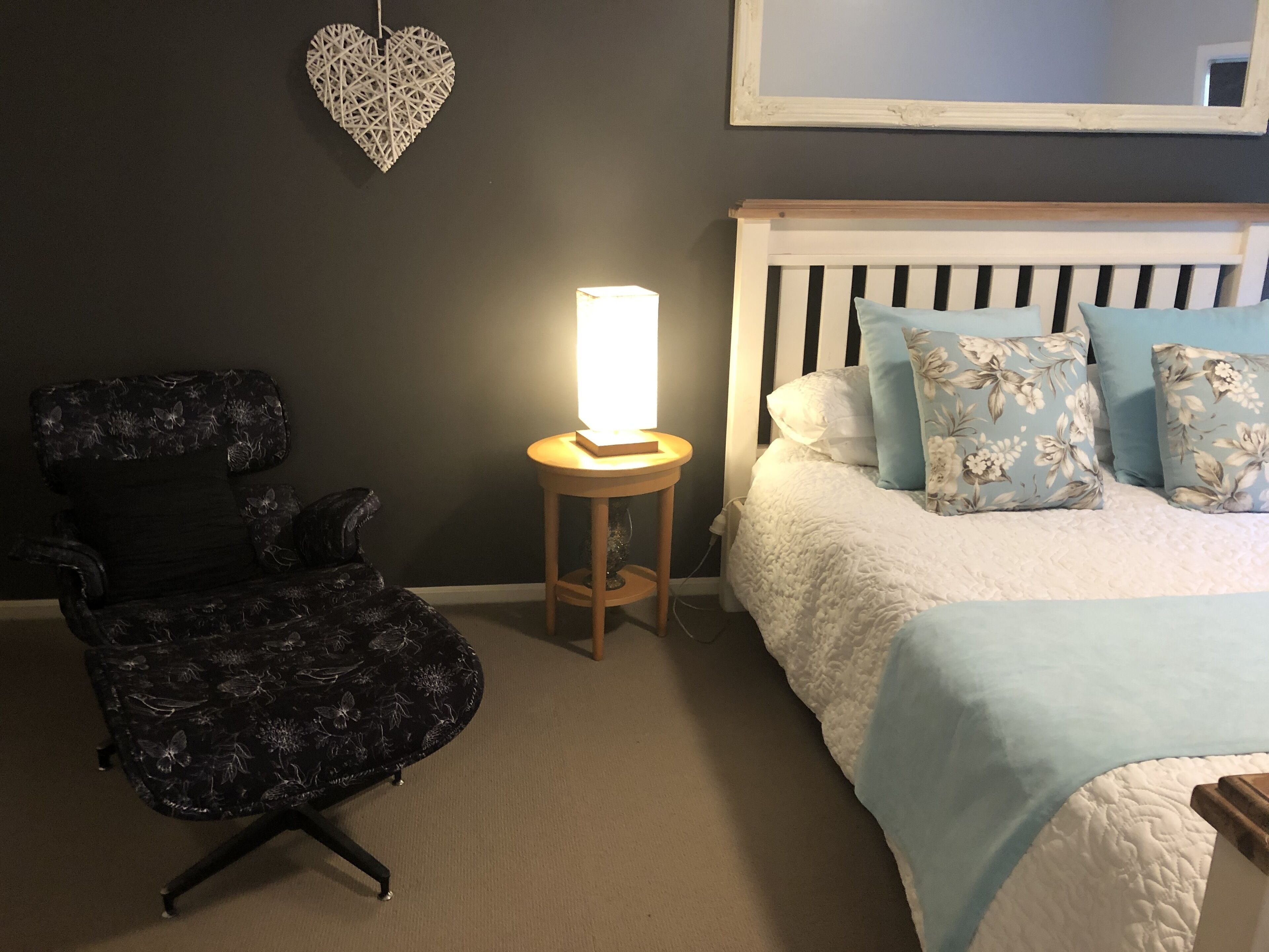 A bedroom with a bed, a chair, a bedside table with a lamp, and a heart-shaped decoration on the wall.