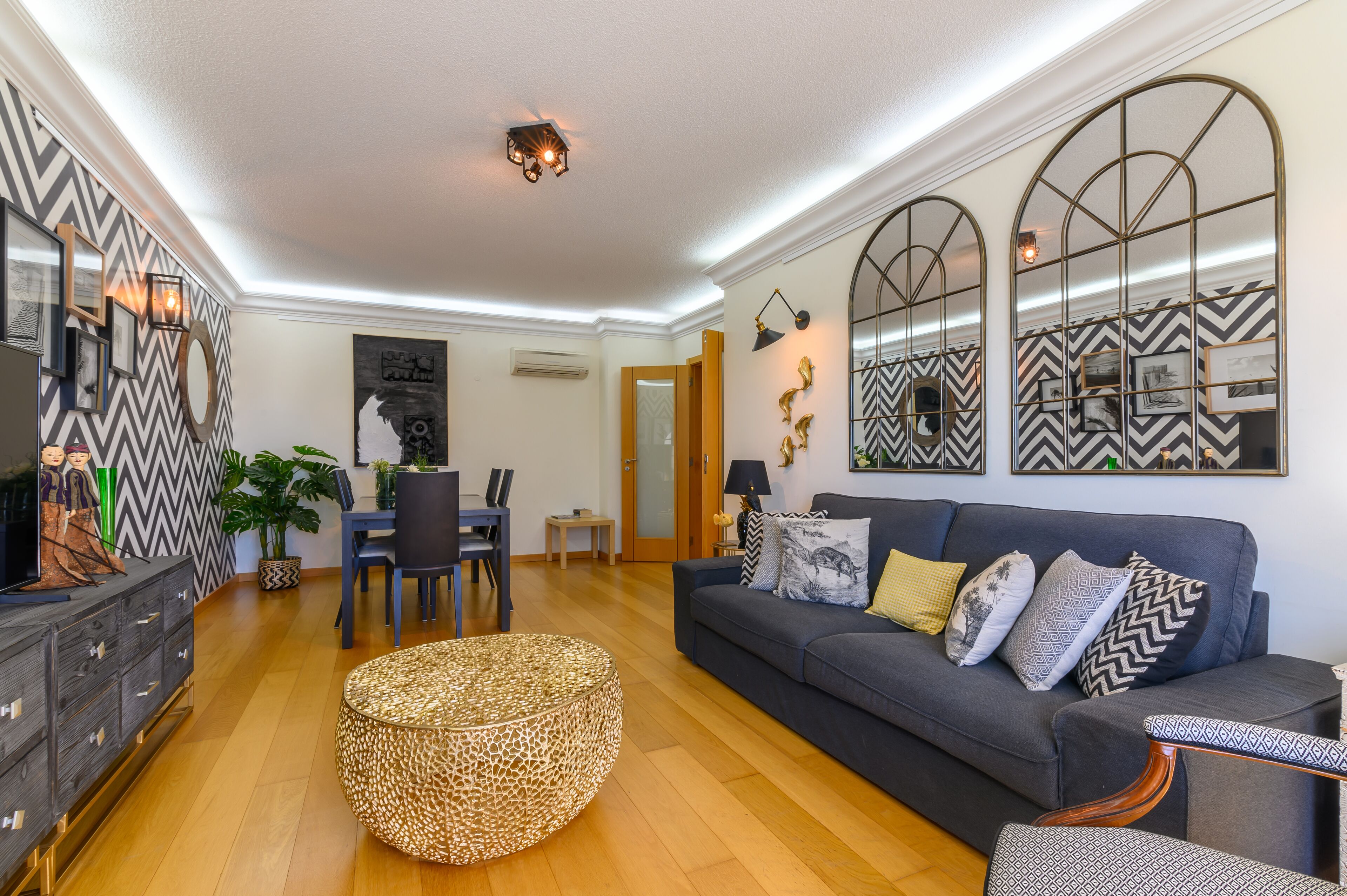 A modern living room with a dark grey sofa, a round woven ottoman, and a dining area with a small table and chairs.