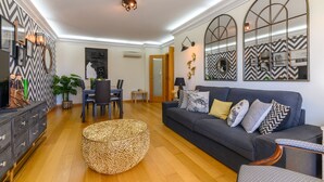 A modern living room with a dark grey sofa, a round woven ottoman, and a dining area with a small table and chairs. - CARCAVELOS BEACH. A wonderful 2 bedroom apartment (Carcavelos)