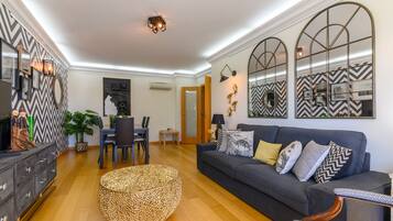 A modern living room with a dark grey sofa, a round woven ottoman, and a dining area with a small table and chairs.