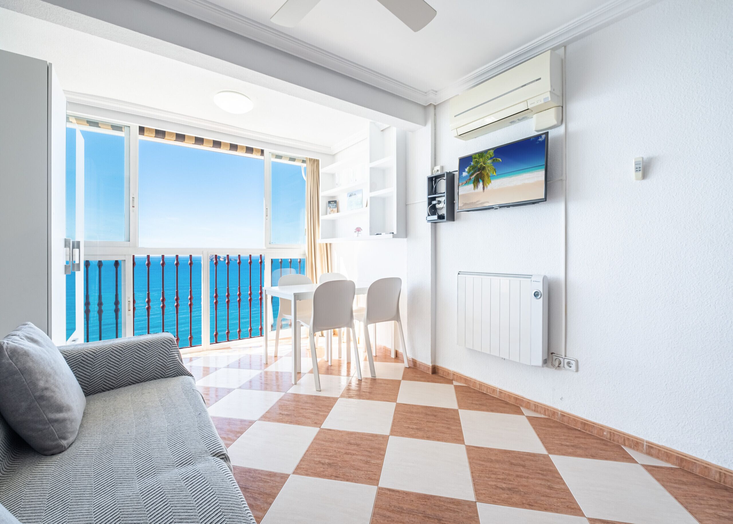 A modern living room with a checkered floor, a sofa, a dining table with chairs, and a view of the sea through large windows.