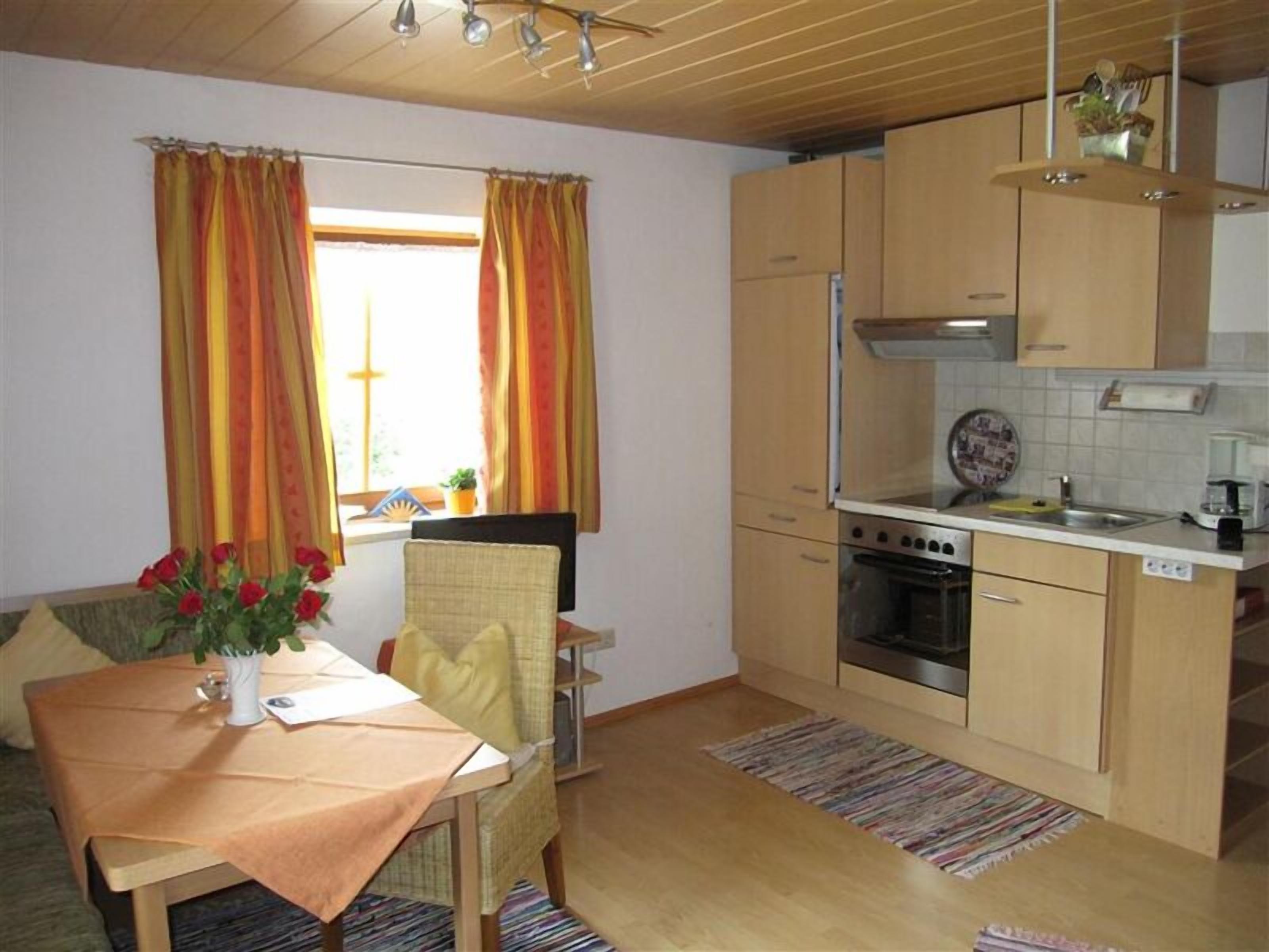 A compact kitchen with a dining area, featuring a table with a vase of flowers, wooden cabinets, and a window with curtains.