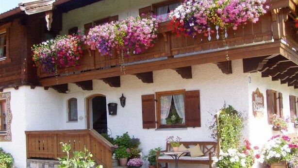 A traditional chalet with wooden balconies adorned with colorful flower boxes, a wooden bench, and a small table with chairs.