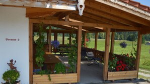 A covered outdoor seating area with wooden benches and a table, surrounded by potted plants and flowers.