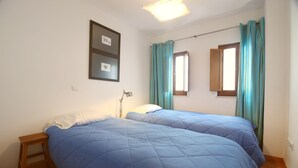 Un dormitorio con dos camas, una mesita de noche, una lámpara y un ventanal con cortinas.
